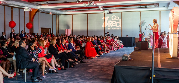 Photo of attendees seated in rows facing female speaker on stage with Go Red for Women logo projected in the background.